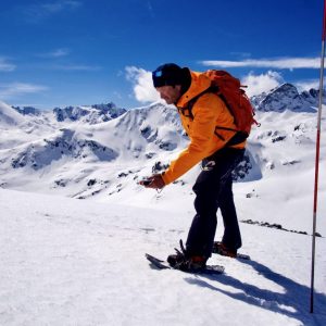 A man wearing orange jacket and backpack holding an avalanche beeper with snowshoes on the mountain