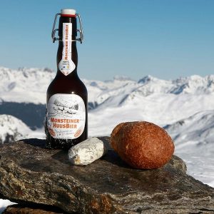 On top of a rock we have two cheeses and a bottle of beer and in the background the mountains full of snow.